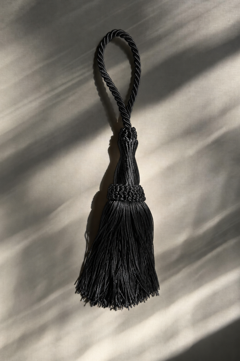 A black braided tassel with a matching hanging loop, photographed against a light neutral background with dramatic diagonal shadows.